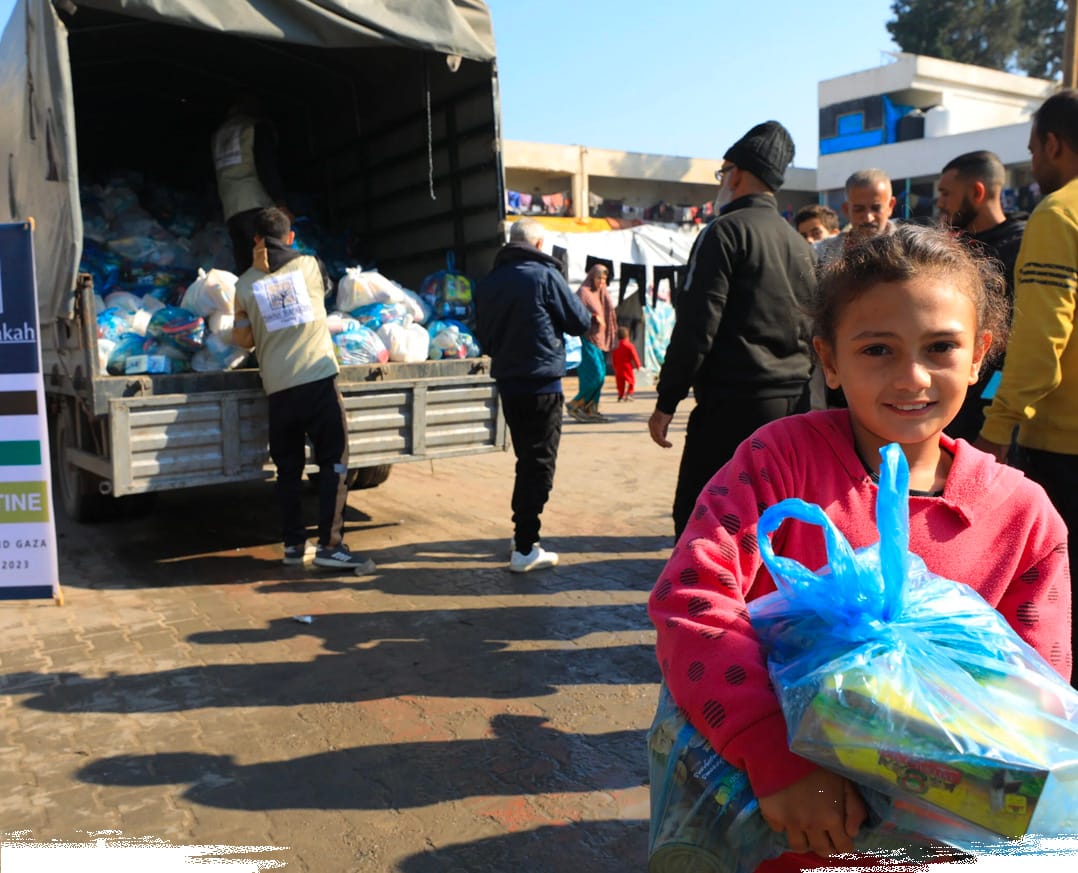 Ein junges Mädchen lächelt, während sie eine blaue Plastiktüte mit Lebensmittelhilfe in der Nähe eines humanitären Hilfslastwagens trägt.