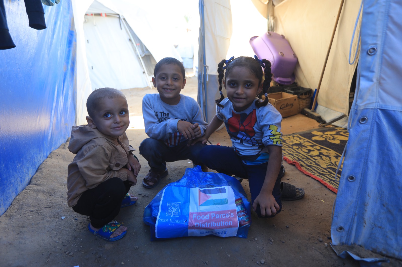 Drei lächelnde Kinder in einem Zelt mit einem blauen Lebensmittelpaket der Barakah Charity; jetzt für Gaza spenden, um Hunger zu lindern.