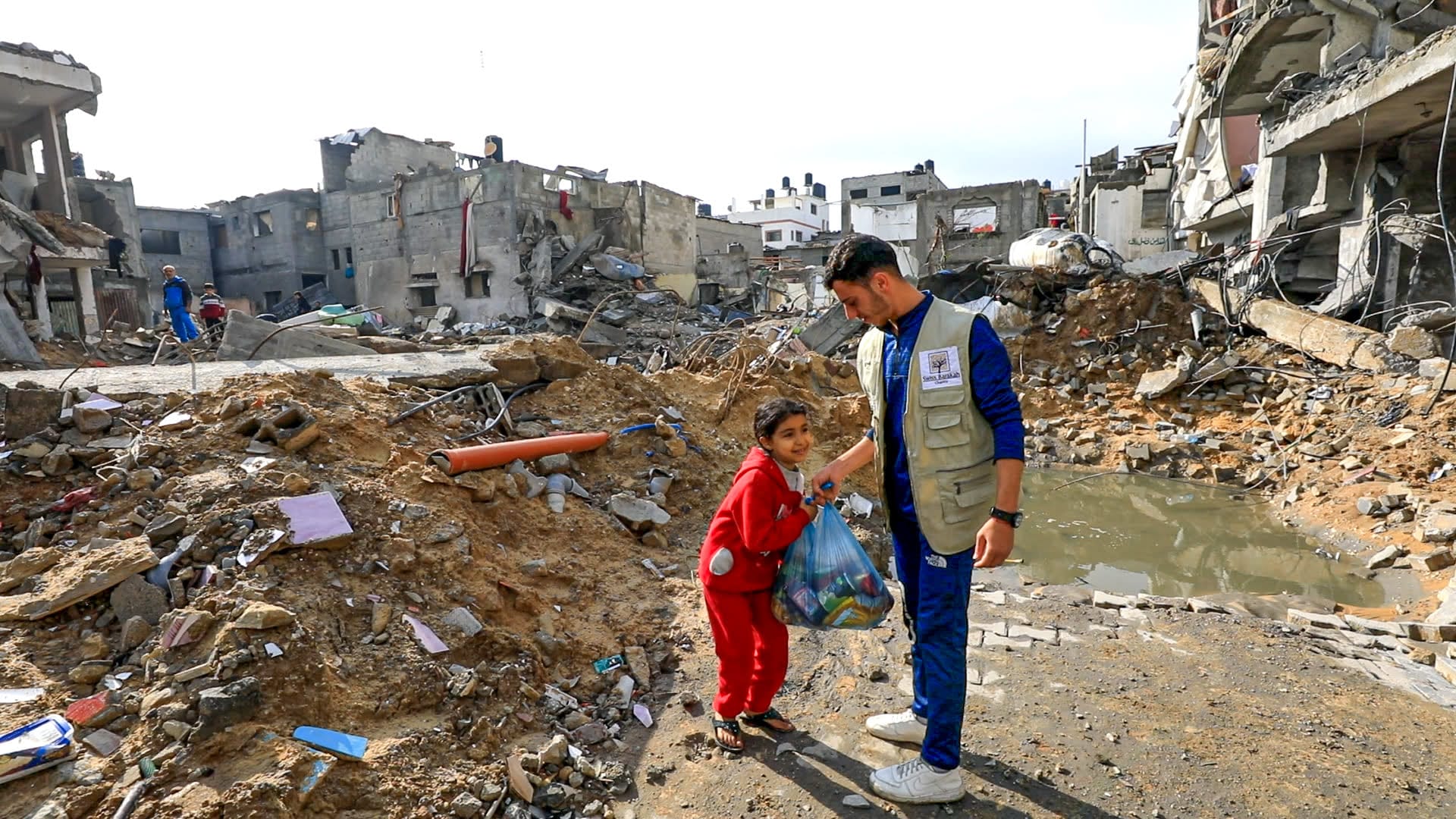 Ein Helfer übergibt einem lächelnden Mädchen in Ruinen eine Lebensmitteltasche. Mit Ihren Spenden für Gaza unterstützen wir Kinder und Familien vor Ort mit überlebenswichtigen Nahrungsmitteln.