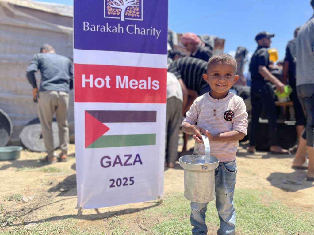 Ein lächelnder Junge mit Metallkübel bei der Verteilung warmer Mahlzeiten; jetzt für Gaza spenden, um die Hungerhilfe zu sichern.