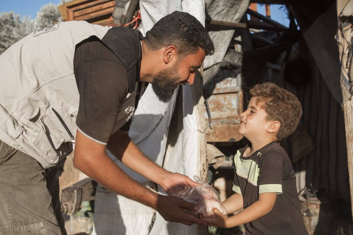 Un operatore manitario che dà del cibo a un bambino. Sotto, opzioni di prezzo per le donazioni qurban in varie regioni (Africa, Yemen, rifugiati palestinesi), con una sezione "Gaza - Prezzo su richiesta".