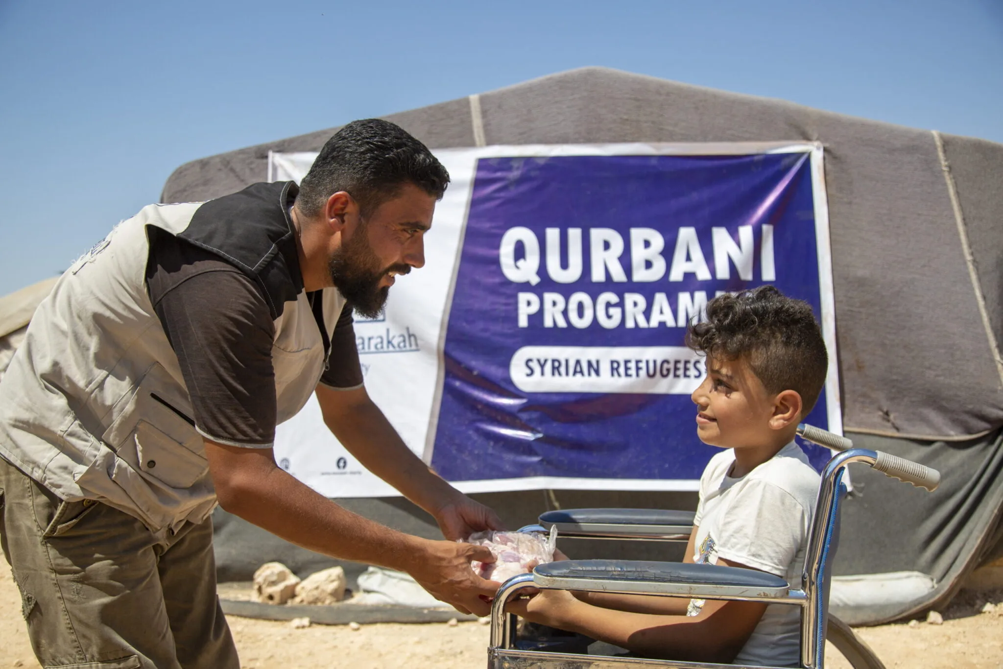 An aid worker handing a package from Kurban donations to a boy in a wheelchair in front of a relief tent.