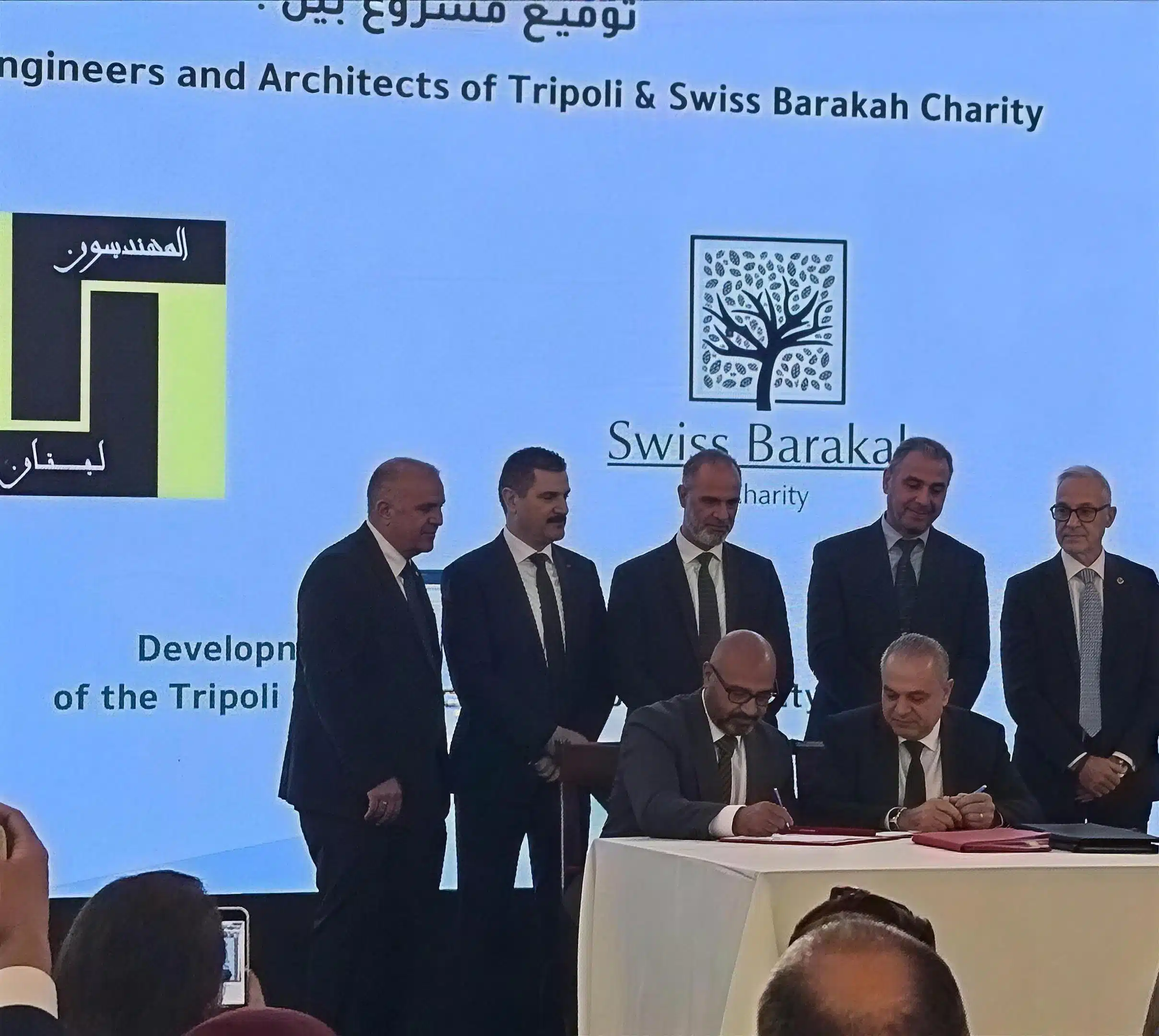 Unterzeichnung der Vereinbarung zwischen der Ingenieur- und Architektenkammer Tripoli und Swiss Barakah Charity, im Hintergrund die Logos beider Organisationen